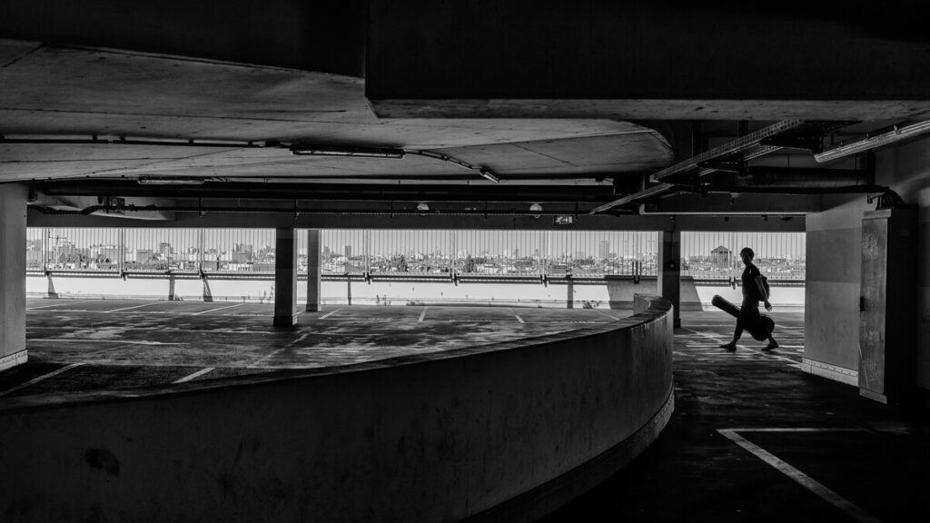 Black and white silhouette of a man carrying a guitar case in a parking garage overlooking the Berlin skyline at Klunkerkranich.