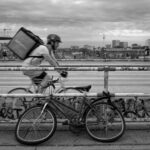 Black and white street photograph of a delivery rider cycling across the Warschauer Brücke in Berlin, with a twisted bicycle in the foreground and the 'Fernsehturm' visible in the urban skyline under a cloudy sky.